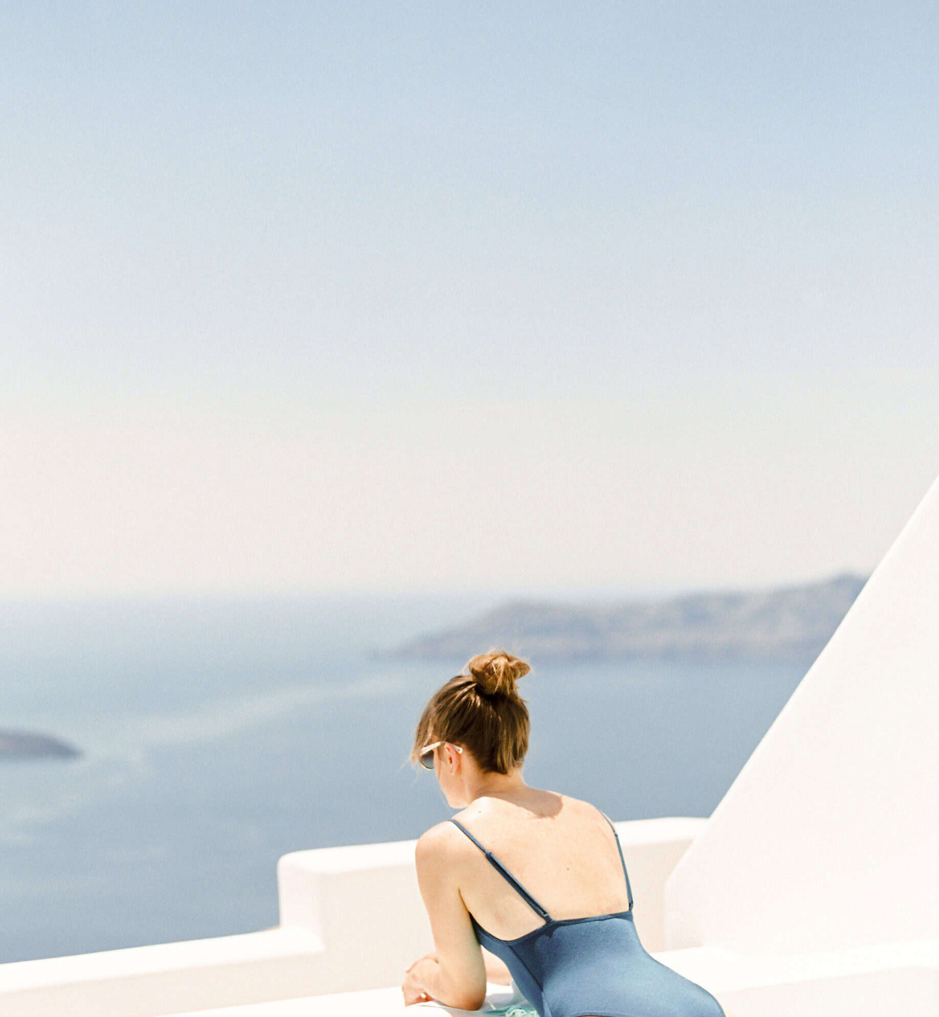 A woman leaning on a white balcony and looking out at the ocean on a sunny day.